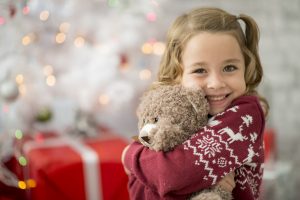 A younggirl is indoors in a living room on Christmas day. She is wearing festive clothing. She is sitting in front of a Christmas tree and hugging her new teddy bear.