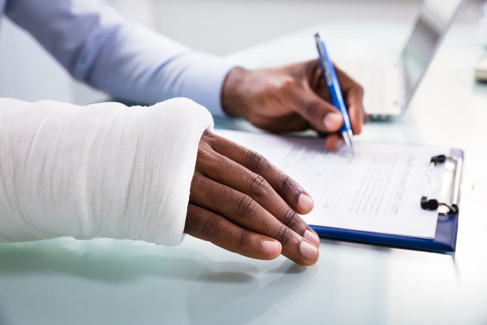 Person with an arm in a cast signing a document next to someone typing on a laptop